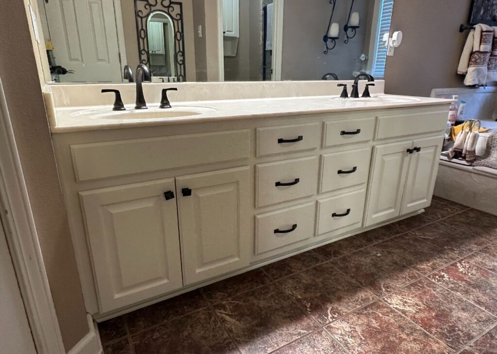A bathroom with a double-sink vanity featuring white cabinets and black handles, brown tiled floor, and a reflection of additional decor in the mirror.