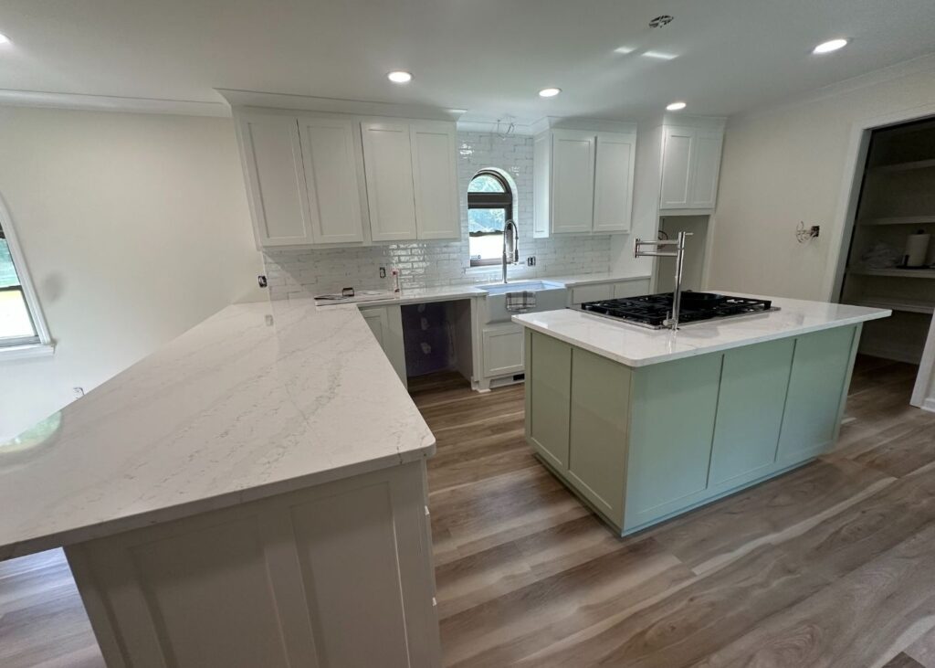 Bright modern kitchen with white cabinetry, light marble countertops, a central island with a cooktop, subway tile backsplash, and wood-look flooring. Natural light enters through an arched window above the sink.