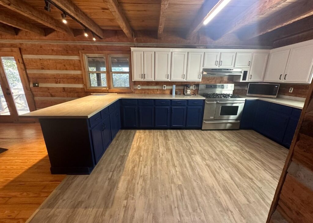A rustic kitchen with exposed wood beams and walls, blue lower cabinets, white upper cabinets, stainless steel appliances, light wood flooring, and a large window letting in natural light.