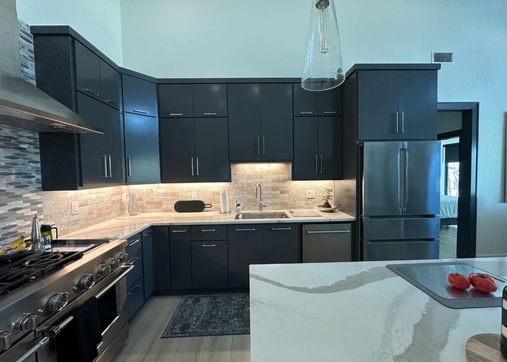Modern kitchen with dark gray cabinets, stainless steel appliances, under-cabinet lighting, a marble countertop island, and a gas stove. A window lets in natural light, and sliced tomatoes are on the island.