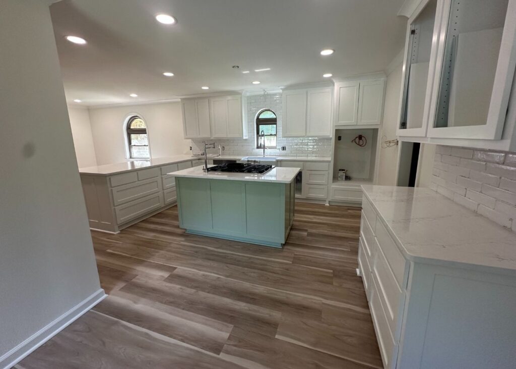 Modern kitchen with white cabinets, marble countertops, light wood flooring, and an island in the center. Two arched windows provide natural light. The walls are light-colored, giving the space a bright and open feel.