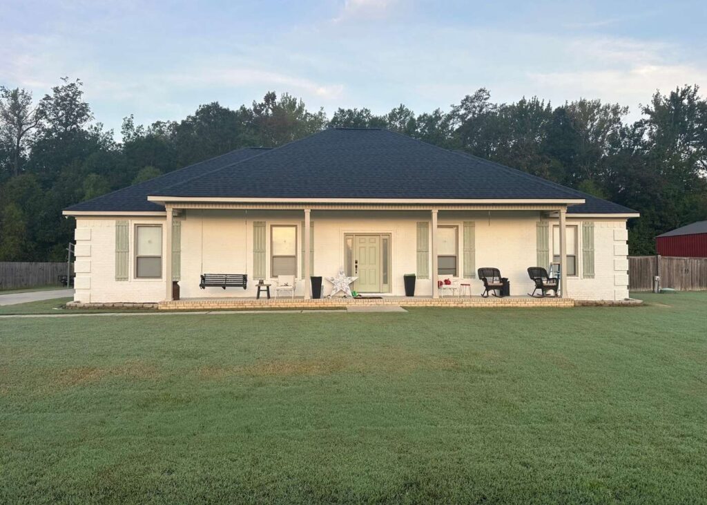 A single-story white house with a dark roof, wide front porch, and columns. The porch has chairs, a bench, and decorations. The house sits on a large grassy yard with trees and a wooden fence in the background.