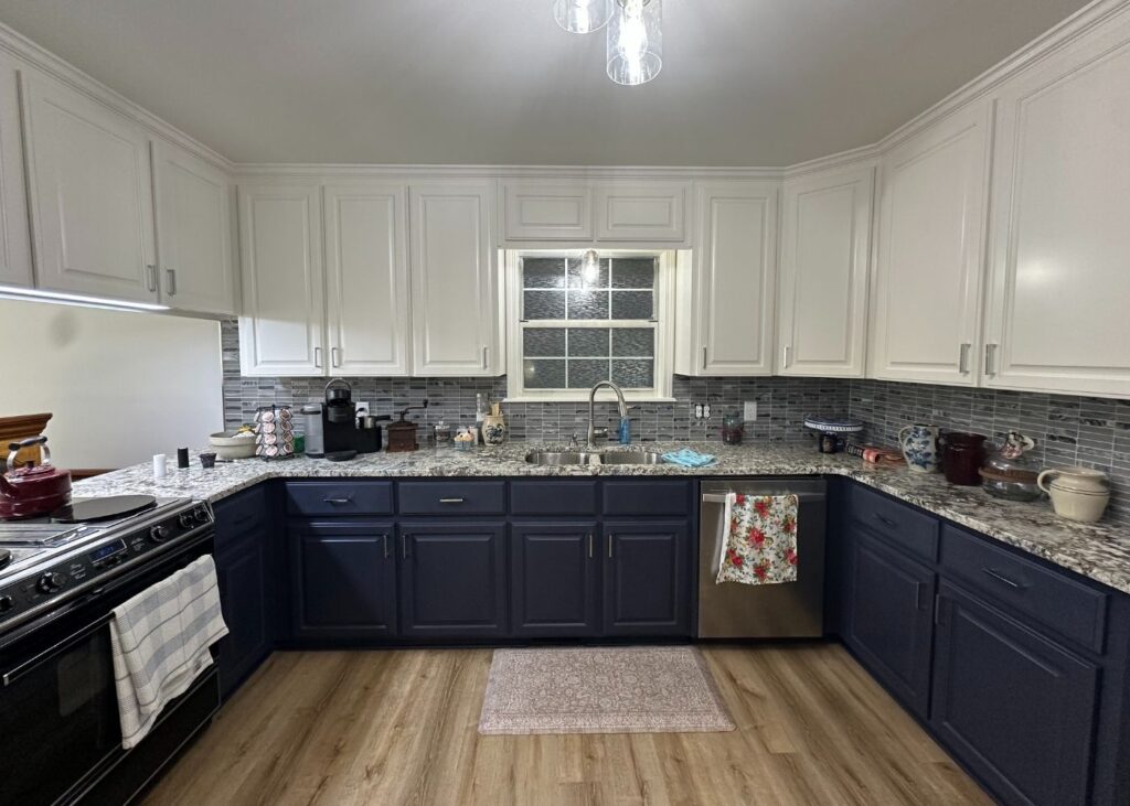 A modern kitchen with white upper cabinets, dark blue lower cabinets, granite countertops, and a gray tile backsplash. A window is above the sink, and various kitchen items are on the counters. A light fixture hangs above.