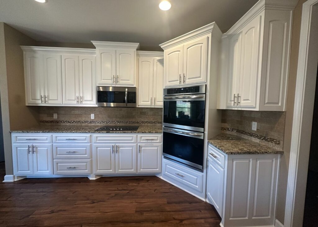 Modern kitchen with white cabinets, granite countertops, brown tile backsplash, built-in double oven, microwave, and wooden floor. The space is well-lit with recessed lighting.