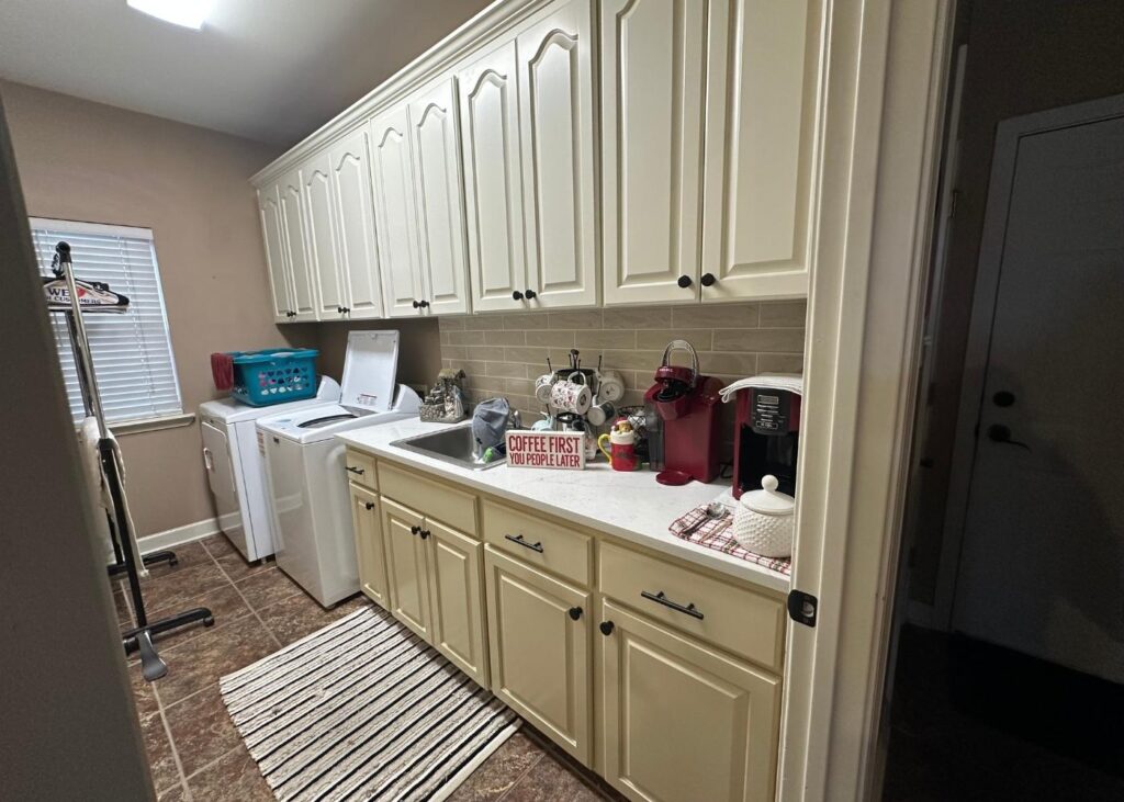A laundry room with white cabinets, a washer, dryer, laundry basket, and countertop with coffee maker, mugs, and a Coffee First, People Later sign. There’s a striped rug on the brown tile floor.