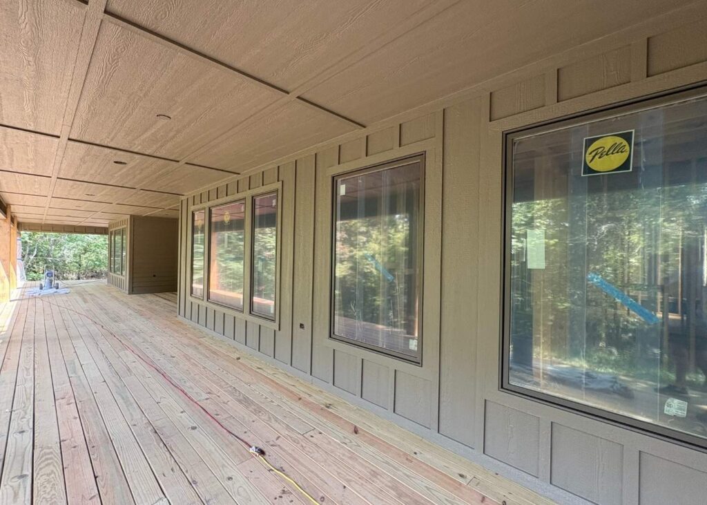 A covered wooden porch with large windows along the side of a tan house, surrounded by trees. The deck is unfinished with construction materials visible in the background.